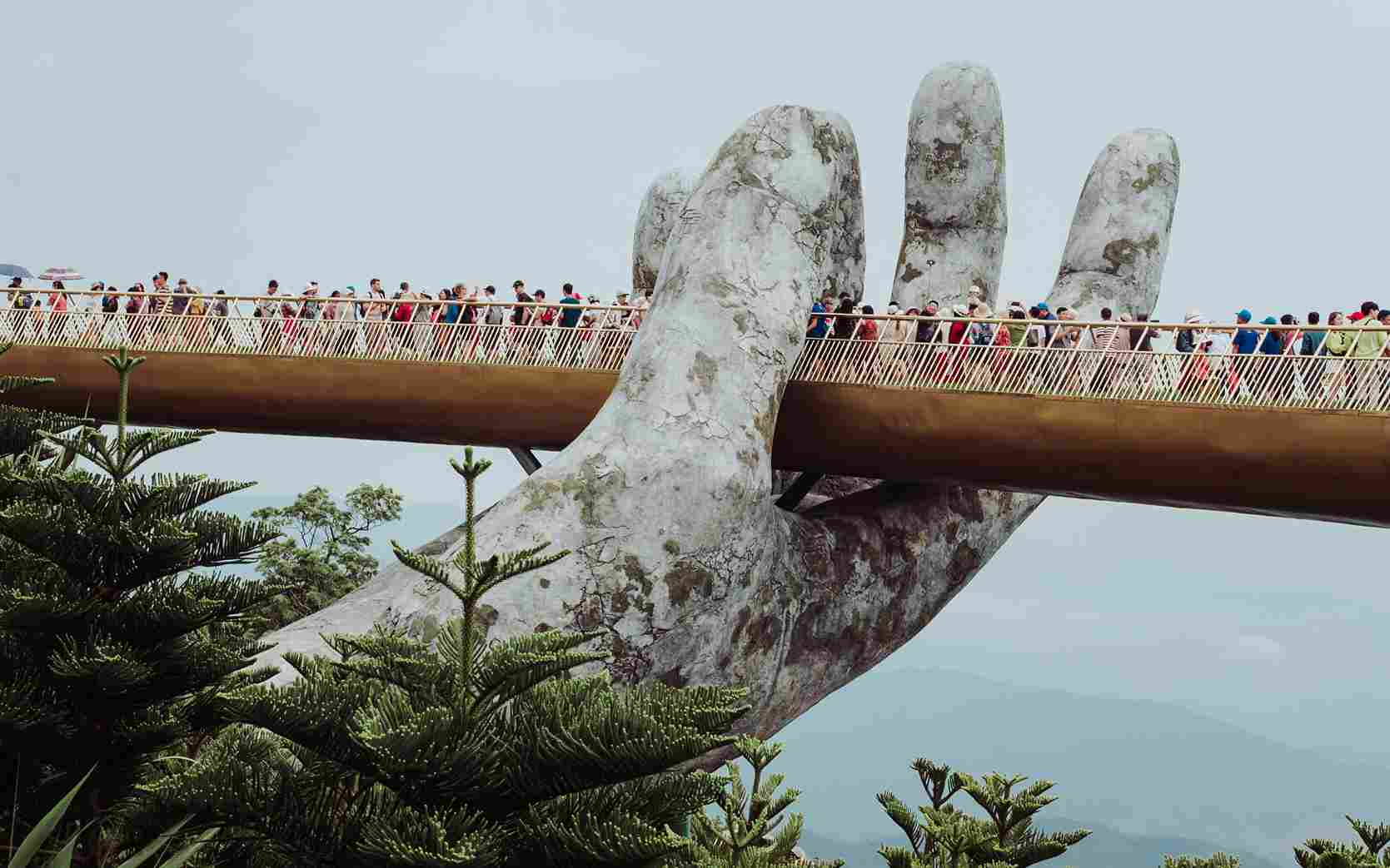 Golden Bridge di Ba Na Hills, Vietnam, sebuah jembatan ikonik yang ditopang oleh dua tangan raksasa batu, dipenuhi wisatawan yang menikmati pemandangan alam pegunungan hijau di sekitarnya.