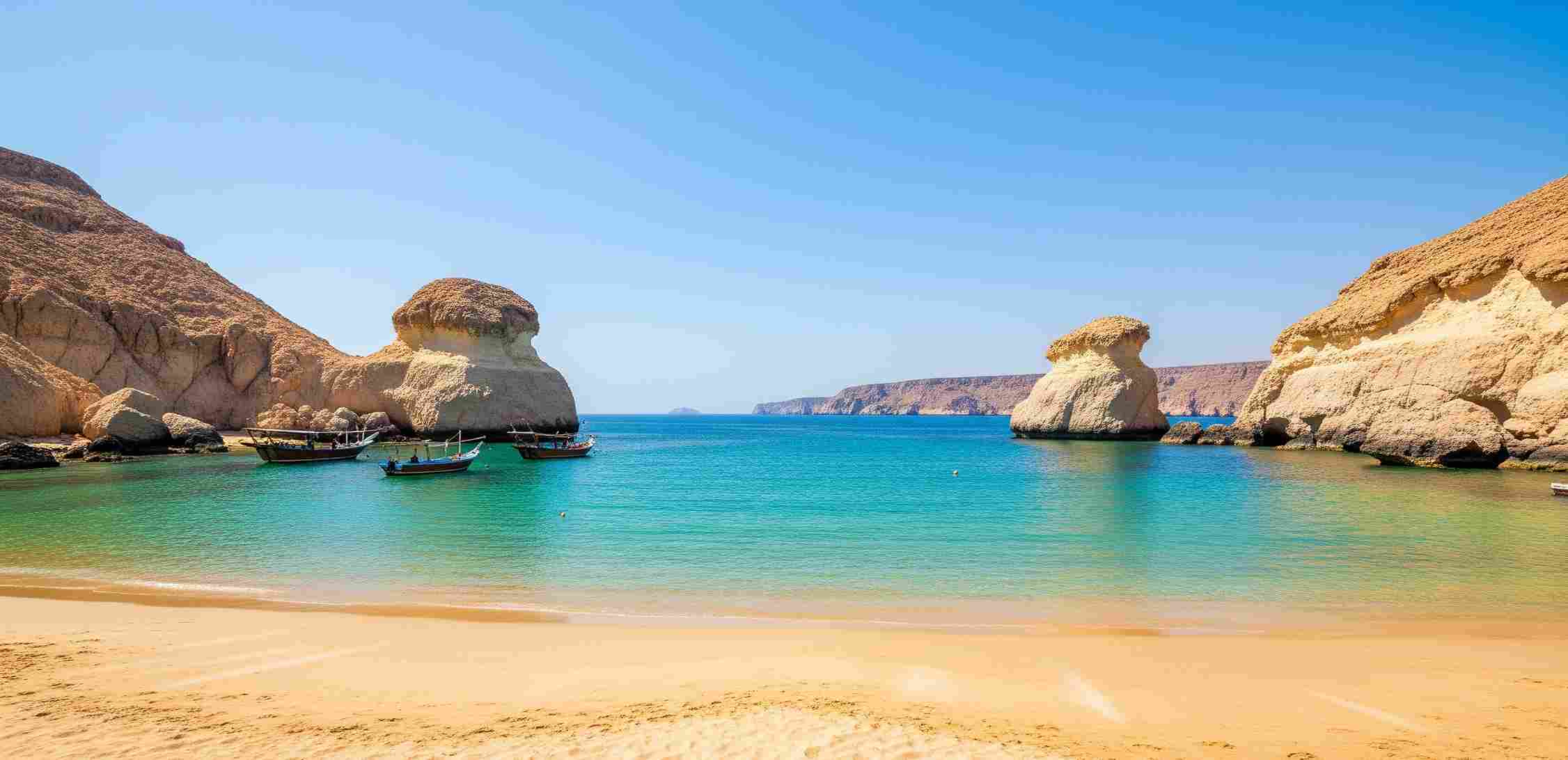 Pemandangan luas Qantab Beach di Oman, menunjukkan pasir keemasan, air pirus yang jernih, dan formasi batuan unik atau tebing yang mengelilingi teluk, menjadikannya salah satu pantai eksotis di Timur Tengah.
