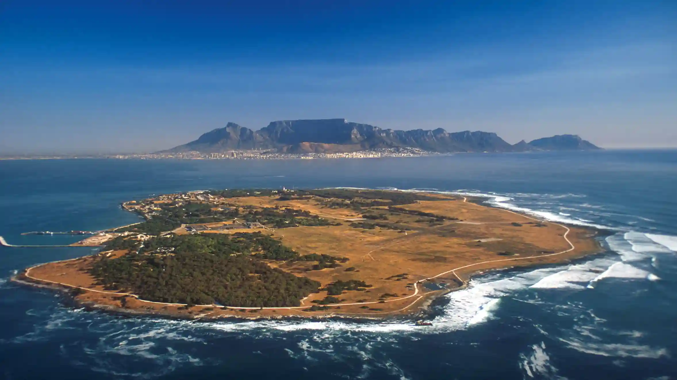 Lanskap Robben Island di Afrika Selatan, menampilkan garis pantai dan medan alami yang terjal, dengan pemandangan Table Mountain dan Cape Town di kejauhan, di bawah langit yang cerah. Pulau ini adalah bagian penting dari wisata di Afrika Selatan yang menawarkan keindahan alam dan sejarah.